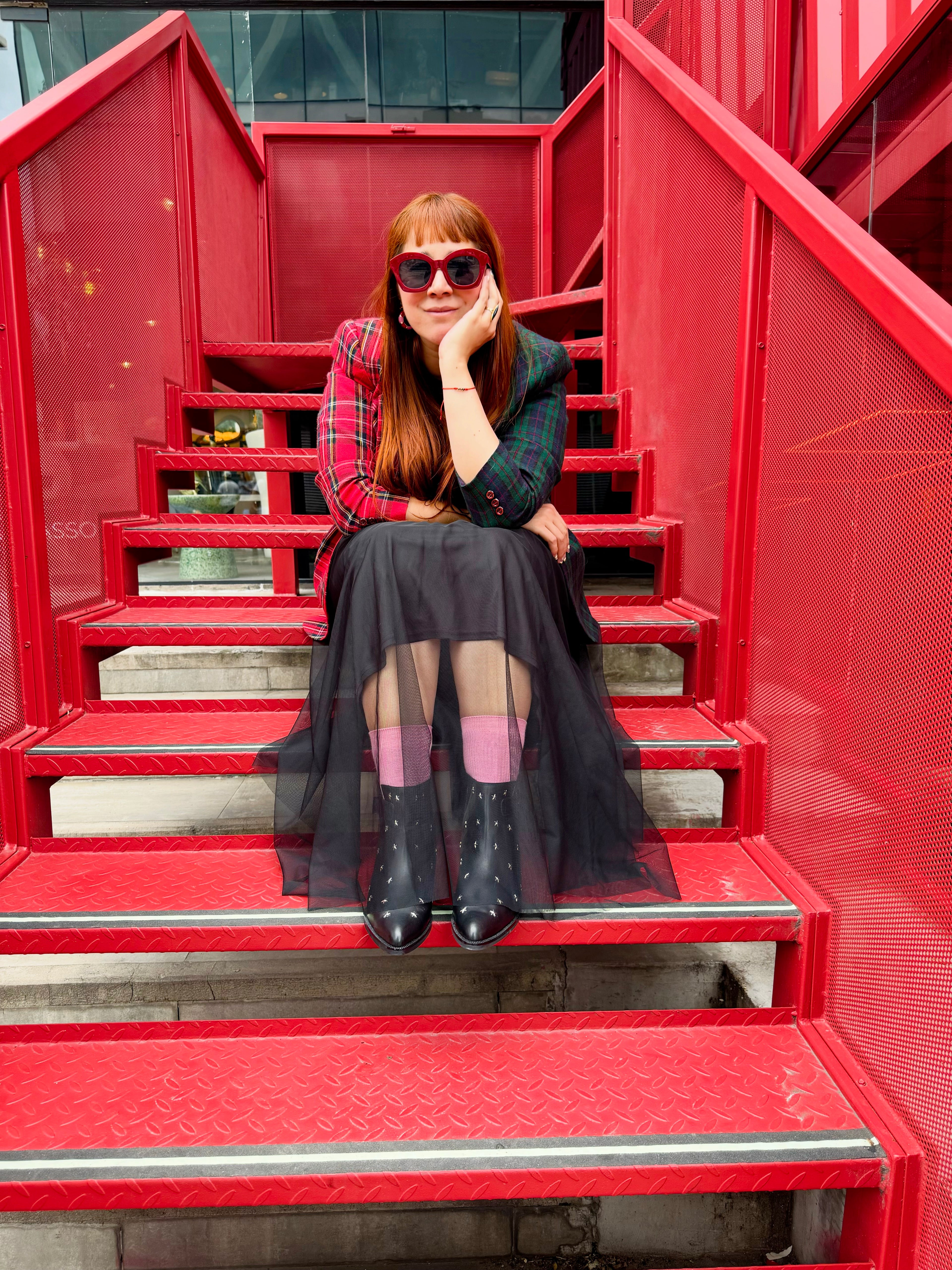 A woman with red hair and sunglasses sits on a set of red metal stairs, wearing a red and green plaid blazer, a dark green patterned top, a sheer black midi skirt, pink tights, and black boots.