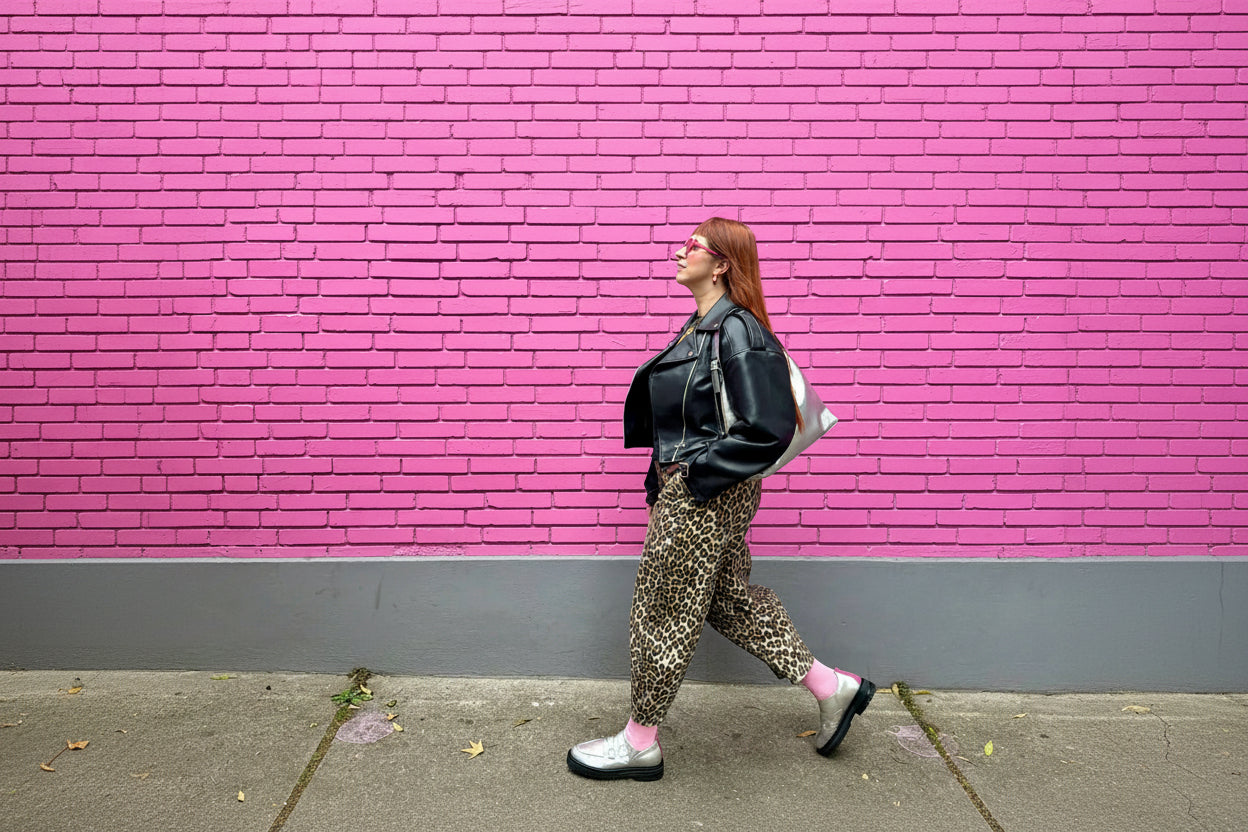 A woman walks in profile against a bright pink brick wall, wearing a black leather jacket, leopard-print pants, pink socks, and black-and-white sneakers.