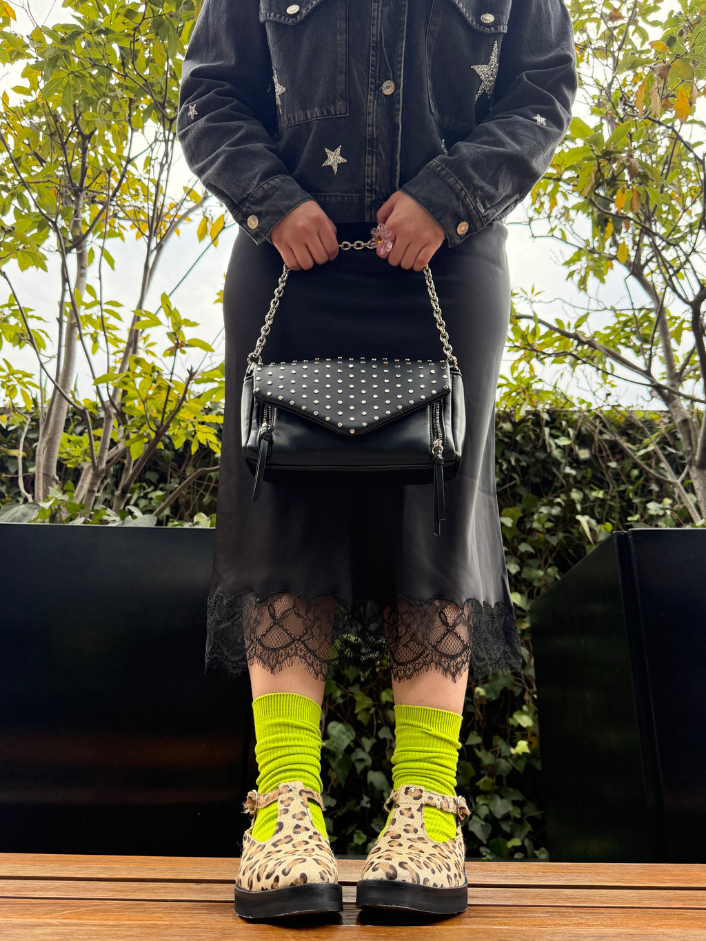 A person wearing a black denim jacket with star details, a black skirt, black lace socks, neon yellow socks, leopard-print platform shoes, and carrying a black studded shoulder bag with a silver chain strap.