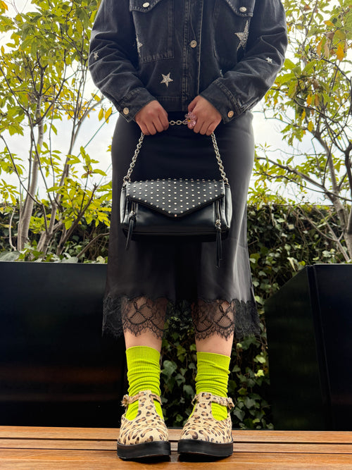 A person wearing a black denim jacket with star details, a black skirt, black lace socks, neon yellow socks, leopard-print platform shoes, and carrying a black studded shoulder bag with a silver chain strap.