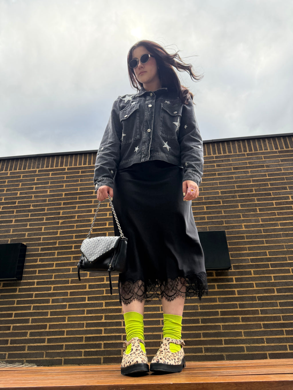 A woman stands outdoors in front of a brick wall wearing a dark gray denim jacket with star patches, a black midi skirt with lace hem, neon yellow socks, and leopard-print Mary Jane shoes. She is holding a black quilted handbag.