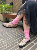 A person sitting outdoors on a brick bench wearing leopard-print Mary Jane shoes with bright pink socks and a black lace-trim skirt.