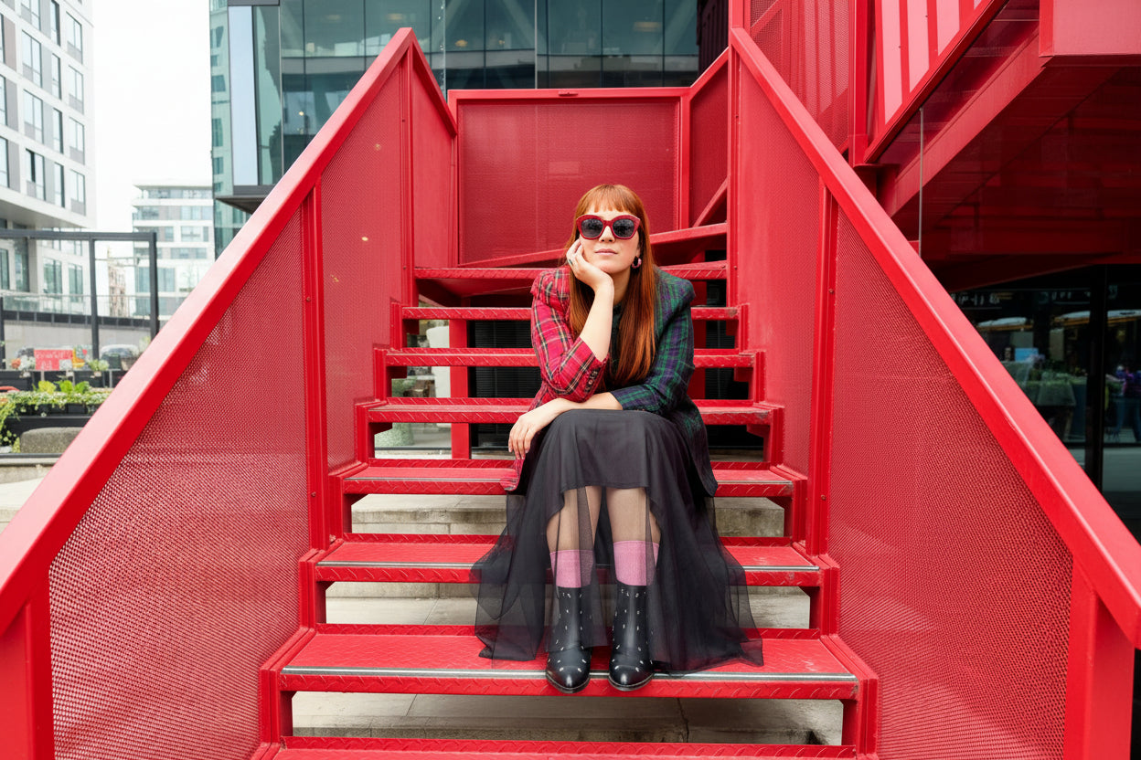 A woman with red hair and sunglasses sits on a set of red metal stairs, wearing a red and green plaid blazer, a dark green patterned top, a sheer black midi skirt, pink tights, and black boots.