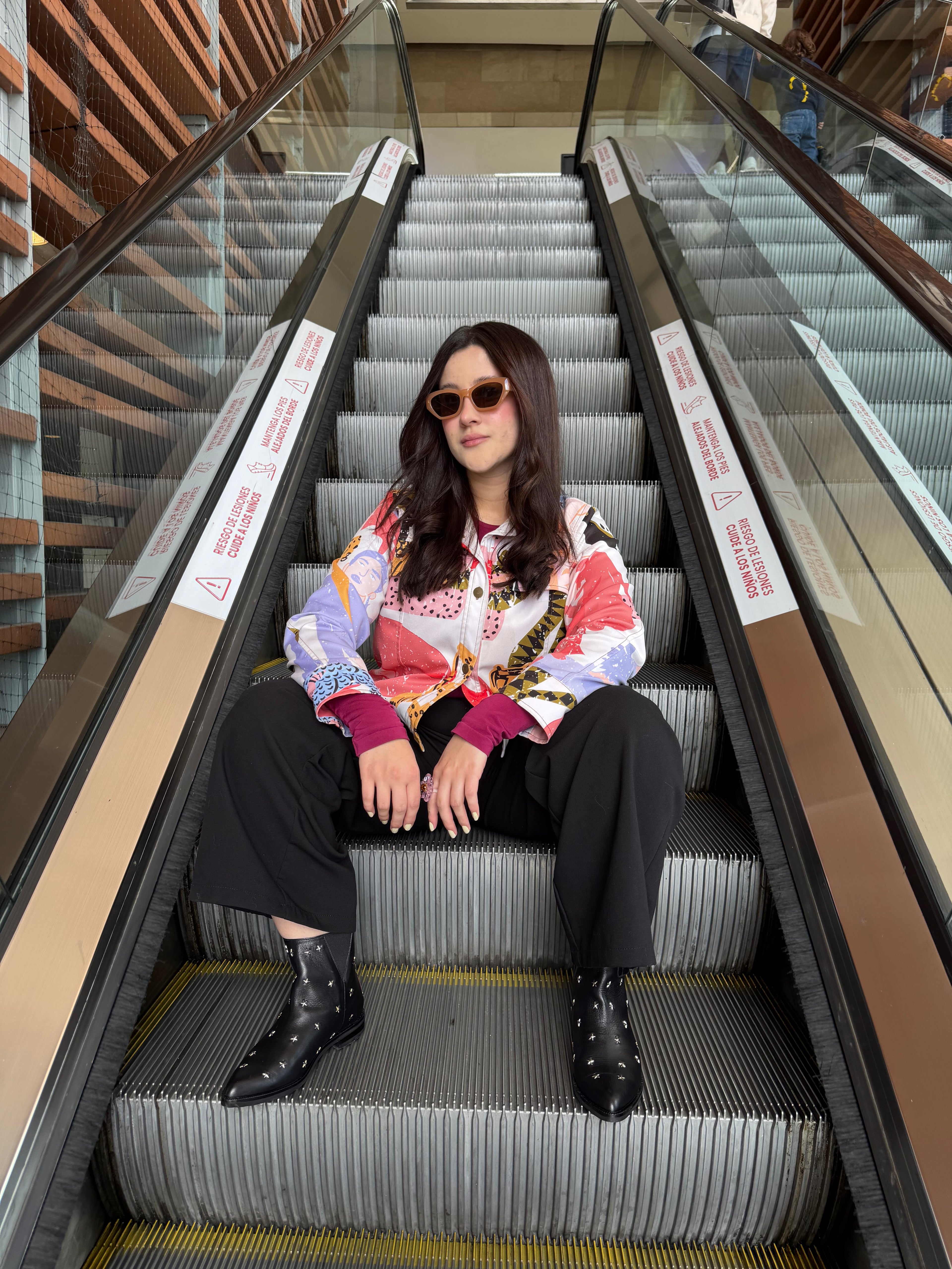 A woman wearing a colorful patterned button-up shirt, black wide-leg trousers, and black studded ankle boots sits on an escalator in a modern indoor setting.